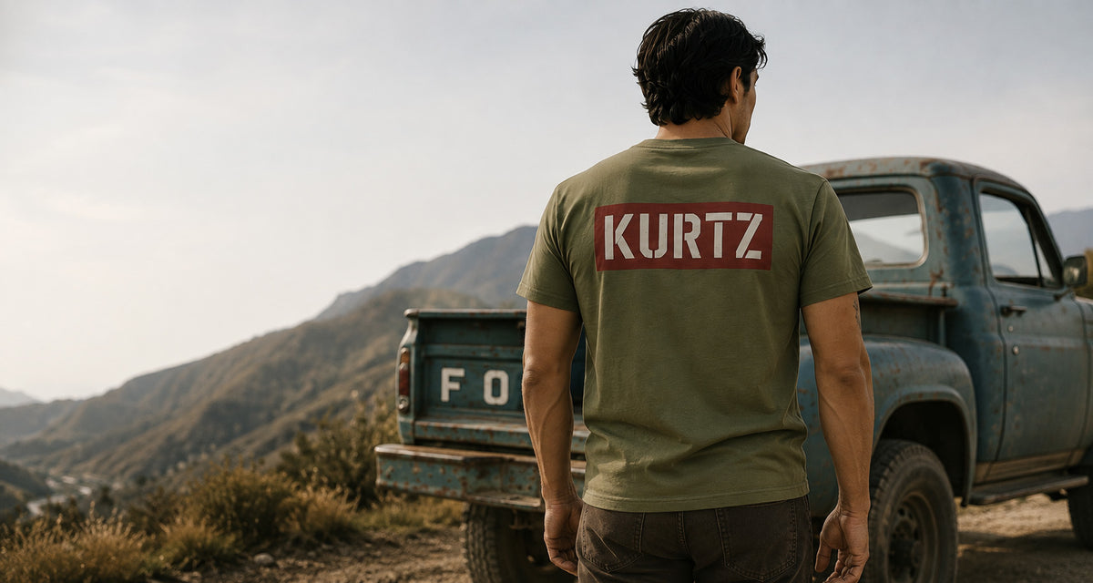 A man viewed from behind wearing an olive green t-shirt with a red rectangular patch on the back that reads 'KURTZ' in white. He is standing outdoors next to a vintage blue pickup truck with mountains in the background.