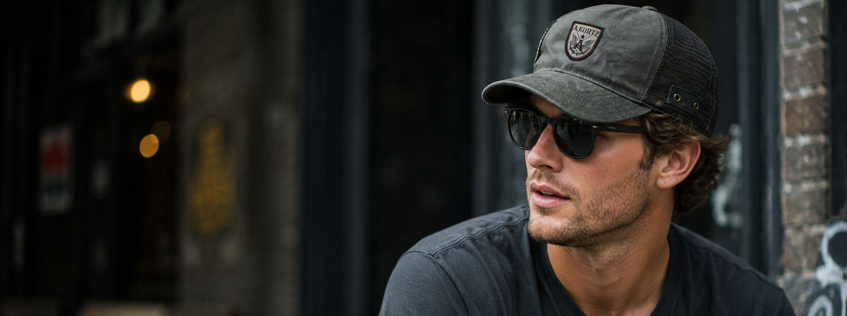 A man wearing a dark gray trucker cap with an A. Kurtz shield logo, black sunglasses, and a dark gray t-shirt, photographed in a dimly lit urban setting.