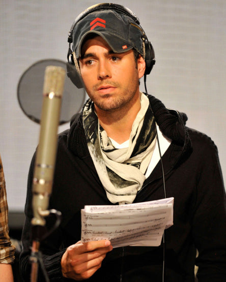 Enrique Iglesias w wojskowej czapce Fritz w kolorze Airborne Navy z czerwonym logo-szewronem, słuchawkach nausznych, czarnej bluzie z kapturem i szarym szaliku, trzymający nuty przed mikrofonem studyjnym.