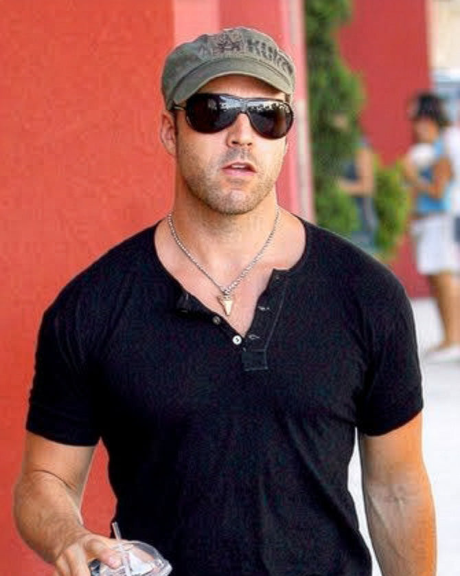 Jeremy Piven w oliwkowozielonej czapce kadetce Kurtz, czarnej koszulce henley i ciemnych okularach przeciwsłonecznych idzie na zewnątrz na tle czerwonej ściany.