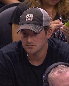 Tony Romo w czapce trucker A. Kurtz Tad, siedzący w zatłoczonej hali lub na arenie.